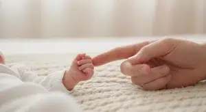 A newborn baby's tiny hand reaching for a parent's finger on a soft cream knit blanket in morning light