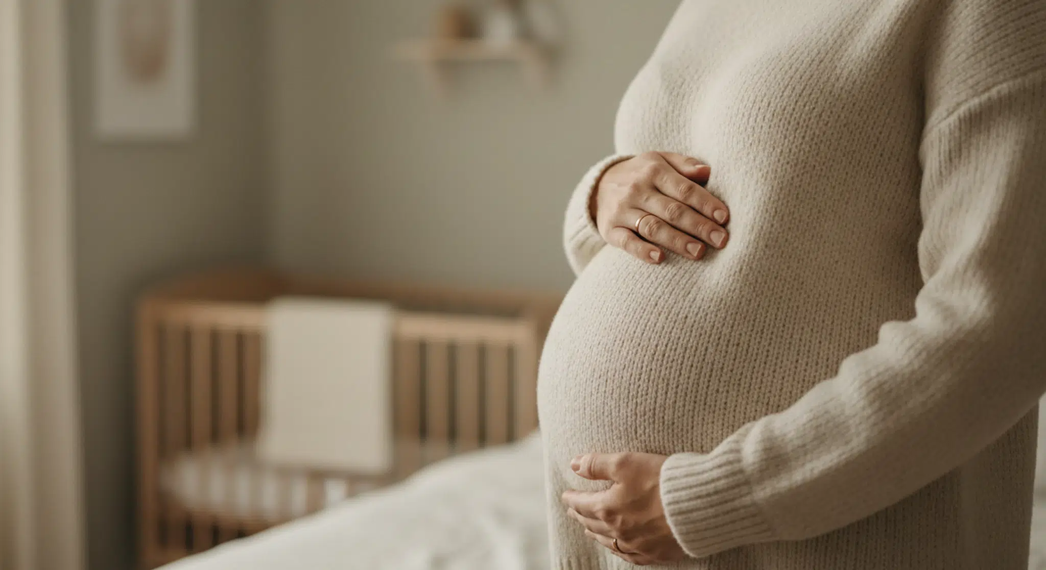 A pregnant woman in a cream knit sweater resting both hands on her belly beside a sunlit window