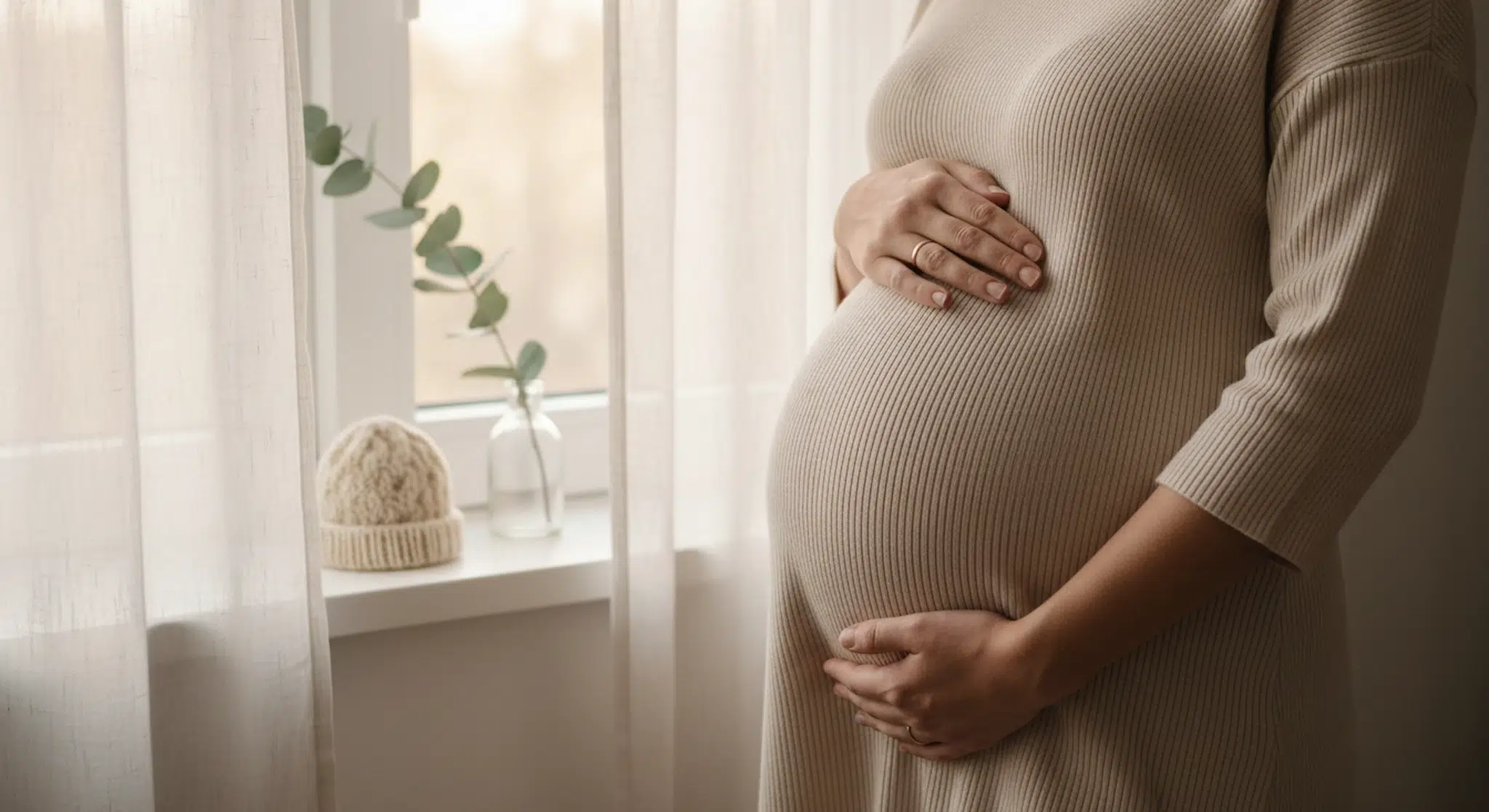 An expectant mother's hands resting gently on her pregnant belly beside a sunlit window, with a small knitted baby hat on the sill