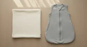 Sleep Sack Vs Swaddle Differences Ages and when to Switch | Betteroo A folded plain swaddle blanket next to a plain sleeveless baby sleep sack laid flat on a changing table