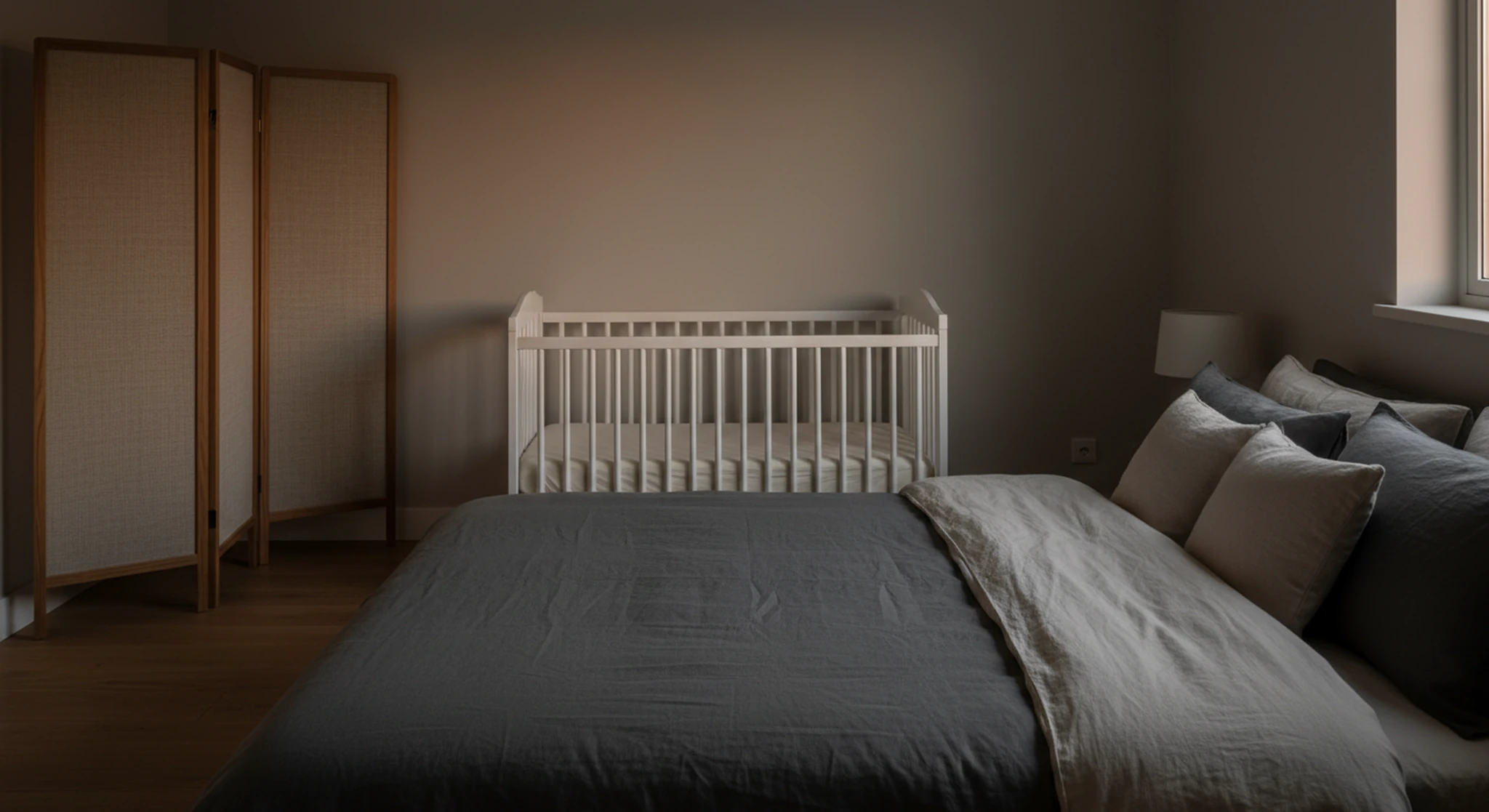 A dim bedroom at dusk with an empty crib on the far side, a folding fabric screen standing apart from it and a made adult bed in the foreground