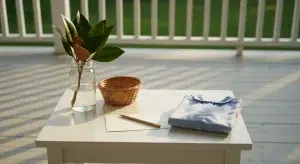 Sunny porch scene with a white table holding a glass jar with plant stems, a small wicker basket, a folded blue shirt, and a pencil on paper on a sunlit deck surface.