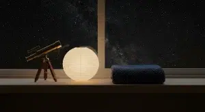 A small brass telescope, a glowing paper lantern and a folded navy dotted baby blanket on a wooden shelf by a starry window