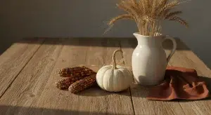A small white pumpkin, dried ears of corn, a ceramic pitcher with wheat stalks and a rust linen napkin on a rustic wooden table