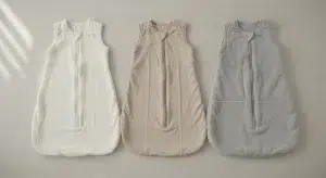 Three plain baby sleep sacks of different thickness laid flat side by side on a light wooden surface