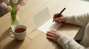 A woman's hands writing in a blank folded card with a pen beside a small vase of pink tulips and a cup of tea